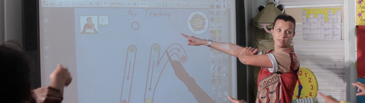 A teacher pointing at a whiteboard screen with the Write2Spell2Read Interactive Whiteboard Program being displayed.